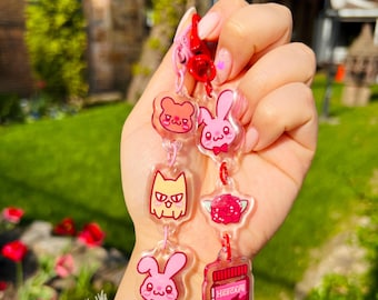 Könnte beinhalten: Eine Sammlung rosafarbener Acryl-Anhänger mit Cartoon-Tieren und einem Glas mit dem Text "Hesgare". Die Anhänger sind durch rote Ketten und einen roten Verschluss verbunden.