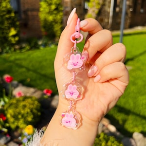 May include: A pink keychain featuring three clear, flower-shaped charms with pink bunny illustrations. The keychain has a pink clasp and connecting rings. The background shows green grass and foliage.