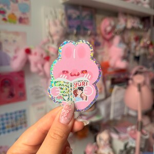 May include: A holographic sticker featuring a pink rabbit character with a rounded head and ears, holding an open book with the text yuri on the cover. The book illustration shows two figures embracing. The sticker has a shimmering, glittery finish.