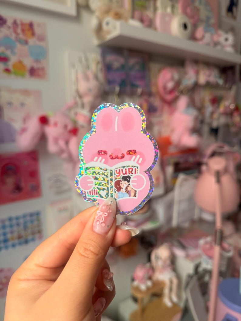 May include: A holographic sticker featuring a pink rabbit character with a rounded head and ears, holding an open book with the text yuri on the cover. The book illustration shows two figures embracing. The sticker has a shimmering, glittery finish.
