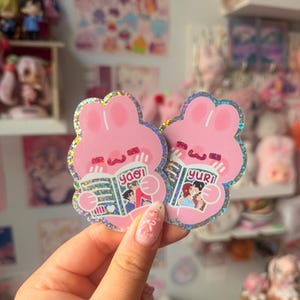 May include: Two holographic die-cut stickers shaped like pink rabbits. Each rabbit holds an open book, one labeled 'yaoi' and the other 'yuri', featuring illustrated characters on the covers. The stickers have a shimmering, glittery finish.