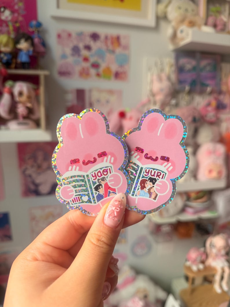 May include: Two holographic die-cut stickers shaped like pink rabbits. Each rabbit holds an open book, one labeled 'yaoi' and the other 'yuri', featuring illustrated characters on the covers. The stickers have a shimmering, glittery finish.