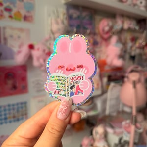 May include: A holographic sticker shaped like a pink rabbit with long ears, holding an open book with the text "yaoi" on the cover. The rabbit has a simple face with two small horizontal lines for eyes and a curved line for a mouth, featuring a glittery finish.