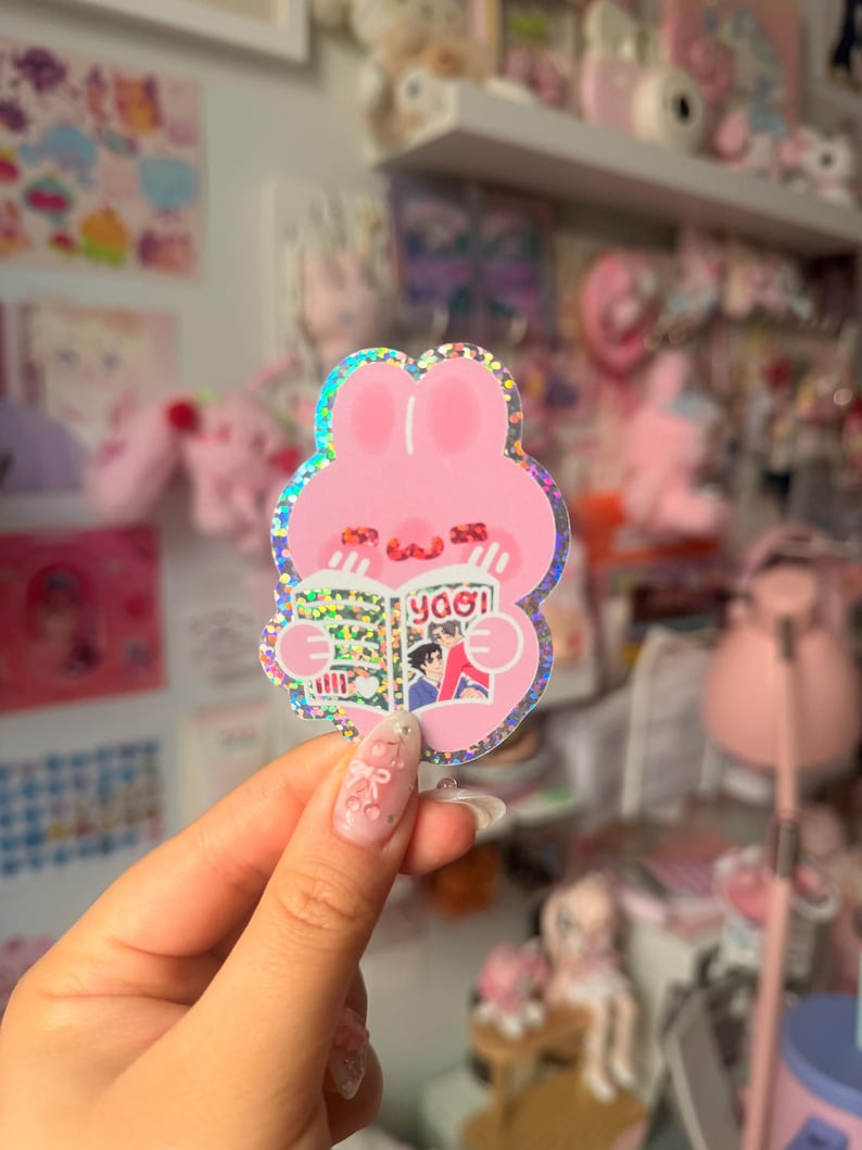 May include: A holographic sticker shaped like a pink rabbit with long ears, holding an open book with the text "yaoi" on the cover. The rabbit has a simple face with two small horizontal lines for eyes and a curved line for a mouth, featuring a glittery finish.