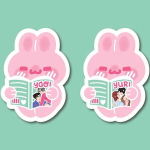 May include: Two die-cut stickers featuring a pink cartoon rabbit character with long ears. Each rabbit holds a light green book. The left sticker shows the text yaoi on the cover with an illustration of two people. The right sticker shows the text yuri.