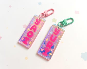 May include: Two rectangular keychains with pink text spelling "HOAY" and "YURI". The keychains have iridescent backgrounds with decorative stars and hearts. One has a pink clasp, the other a green clasp.