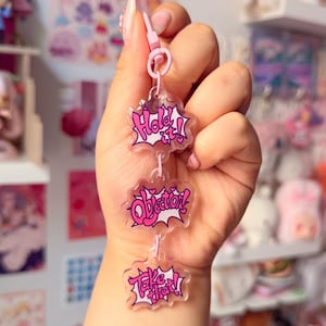 Pode incluir: Um porta-chaves de acrílico transparente com três pingentes empilhados em forma de nuvem, conectados por anéis cor-de-rosa. Cada pingente exibe texto rosa com contornos brancos lendo Hold it!, Objection! e Take that! em estilo banda desenhada.
