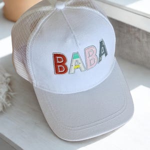 May include: A white trucker hat featuring a mesh back and a solid front panel. The word BABA is embroidered on the front in colorful, block-style letters. The letter B is red, the A features a green and yellow pattern, the second B is light pink, and the final A is dark green.