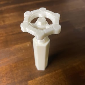 May include: A cream-colored, 3D-printed valve handle with a hexagonal base and a circular top with a star-like design. The handle is designed to be screwed into a fitting. The background is a dark brown wooden surface.