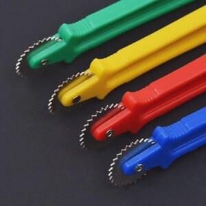 Puede incluir: Cuatro herramientas de plástico de colores con cuchillas dentadas. Los mangos son verdes, amarillos, rojos y azules. Las cuchillas son de metal. Las herramientas están dispuestas en diagonal sobre un fondo oscuro. Estas herramientas probablemente se utilizan para cortar o marcar.