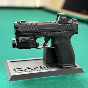 May include: A black Canik Mete MC9 semi-automatic pistol featuring a Holosun red dot optic, a mounted tactical light, and a gold-coloured trigger. The firearm is displayed on a black stand embossed with the text Canik Superior Firearms.