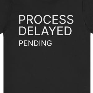 May include: Black t-shirt featuring the white text "PROCESS DELAYED PENDING". The short-sleeved shirt has a crew neck. The text is centrally positioned on the front of the garment.