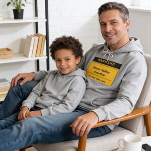 Op de afbeelding: Een vader en kind zitten samen, beiden gekleed in grijze hoodies en een blauwe jeans. De hoodie van de vader heeft een geel en zwart waarschuwingslabel met de tekst "Brain Buffer Overflow: Too many tabs open."