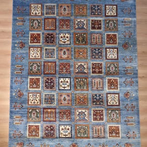 Könnte beinhalten: Ein rechteckiger blauer Teppich mit einem sich wiederholenden Muster aus Quadraten in Braun-, Beige- und Grüntönen. Der Teppich hat ein detailliertes, traditionelles Design mit Fransen am oberen Rand. Der Teppich ist wahrscheinlich aus Wolle oder einem ähnlichen Material.
