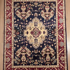 Può includere: Un tappeto rettangolare blu scuro con un intricato motivo floreale. Il tappeto presenta un medaglione centrale color crema e bordeaux, circondato da motivi floreali color crema, bordeaux e oro. Un bordo bordeaux incornicia il disegno.