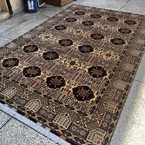 May include: A rectangular area rug featuring a repeating pattern of dark maroon floral medallions set against a cream-colored background. The rug includes intricate geometric borders in shades of brown, tan, and black, finished with white fringe.
