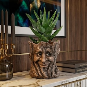 May include: A decorative aloe vera plant in a tree-face planter. The brown planter has a smiling face carved into it. A gold candelabra with dark candles sits to the left. The planter is on a marble surface.