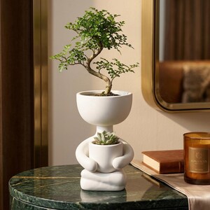 May include: A matte white ceramic planter shaped like a person sitting in a cross-legged position. The figure holds a smaller cylindrical pot containing a succulent, while a small green bonsai tree grows from the larger head-shaped top section.