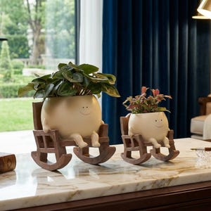 May include: Two whimsical cream-colored planters with smiling faces, each holding a plant. They are seated in brown rocking chairs. One planter has green leafy plants, the other has red and green plants. The planters are on a marble surface.