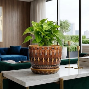 Op de afbeelding: Een bruine keramische plantenpot met bijpassende schotel, gevuld met weelderig groen gebladerte. De pot heeft een herhalend patroon van geometrische vormen in een lichtere bruine kleur. Hij staat op een wit marmeren oppervlak, met gouden kandelaars op de achtergrond.
