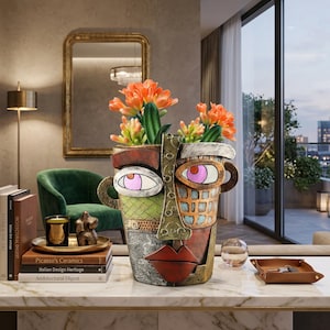 May include: A decorative face-shaped vase with orange flowers. The vase has a colorful, abstract design with large eyes, a red mouth, and a textured surface. It is placed on a marble table in a living room setting.