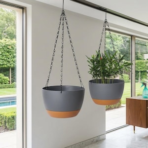 Op de afbeelding: Twee hangende plantenpotten met een grijs bovenstuk en een oranje-bruine basis. Ze hangen aan zwarte kettingen. Een van de potten bevat groen. De potten hangen voor een groot raam.