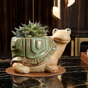 May include: A ceramic turtle-shaped planter with a green and tan glaze, holding a green succulent plant. The turtle has a smiling face and detailed shell design. The planter sits on a round, brown leather coaster.