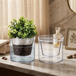 May include: Two clear, cylindrical, double-walled glass planters. One planter contains a small green leafy plant rooted in dark soil topped with white pebbles and water. The other planter is empty, showing the inner and outer glass layers.