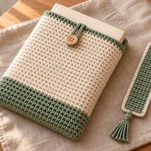 Può includere: Una custodia per libri all'uncinetto color crema e verde salvia con chiusura a bottone in legno, contenente un libro, accompagnata da un segnalibro rettangolare all'uncinetto abbinato con centro verde salvia, bordo crema e nappa verde salvia.