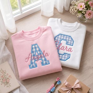 May include: A light pink sweatshirt featuring a blue and white gingham letter A with the name Angela embroidered in pink, and a white sweatshirt featuring a blue and white gingham letter C with the name Clara embroidered in pink.