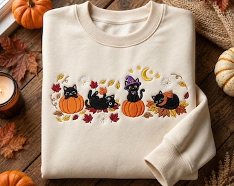 May include: A cream-coloured crewneck sweatshirt featuring an embroidered design of four black cats. The cats are depicted playing with orange pumpkins, wearing a purple witch hat, and resting on colourful autumn leaves with a yellow crescent moon.