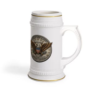 May include: White ceramic beer stein with a gold rim and handle. The stein features a circular emblem with an eagle, the text "IN GOD WE TRUST", and the years 1776-2026. The stein has a textured design around the base and top.