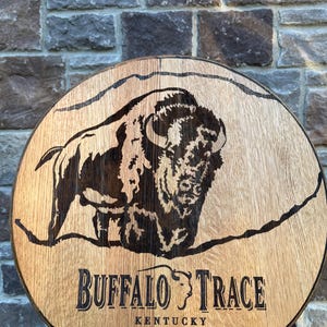 Puede incluir: Un letrero de madera circular que presenta una ilustración oscura quemada de un bisonte. Debajo de la imagen, el texto dice "BUFFALO TRACE" con "KENTUCKY STRAIGHT BOURBON WHISKEY" impreso debajo en una fuente serif negra y clara.