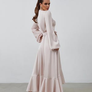 May include: A pale pink maxi dress with long sleeves and a matching belt. The dress has a high neckline and flared sleeves. The skirt features a ruffled hem. The model is wearing high heels.