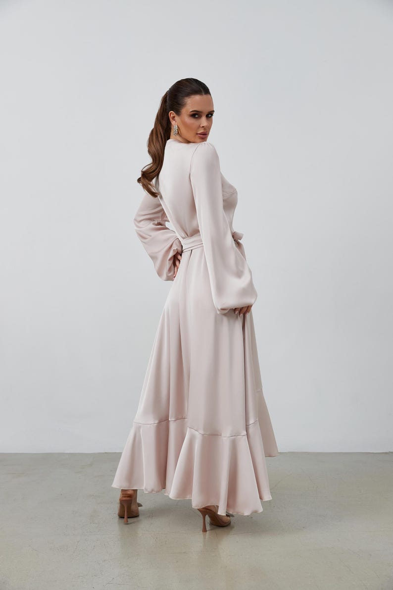 May include: A pale pink maxi dress with long sleeves and a matching belt. The dress has a high neckline and flared sleeves. The skirt features a ruffled hem. The model is wearing high heels.