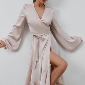 May include: A light beige wrap dress with long, flowing sleeves and a high slit. The dress features a V-neckline, a tie at the waist, and a ruffled hem. The dress is made of a silky material.