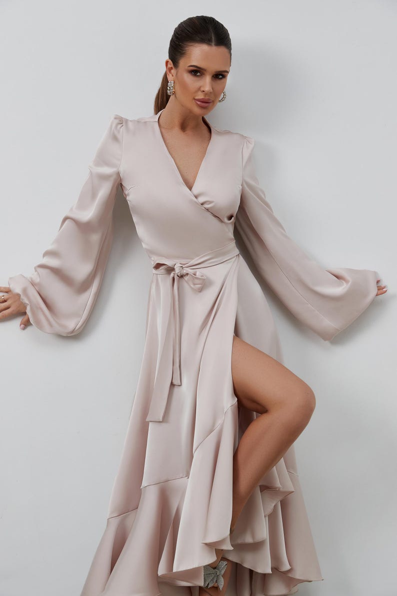 May include: A light beige wrap dress with long, flowing sleeves and a high slit. The dress features a V-neckline, a tie at the waist, and a ruffled hem. The dress is made of a silky material.
