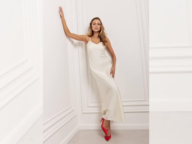 May include: A cream-coloured slip dress with spaghetti straps and a flowing silhouette. The dress is paired with bright red high heels. The model is leaning against a white wall with decorative moulding, creating a classic and elegant look.