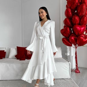 May include: A white wrap dress with long sleeves and a ruffled hem. The dress is tied at the waist with a matching belt. The model is wearing white heels with ankle straps. A bouquet of red heart-shaped balloons is in the background.