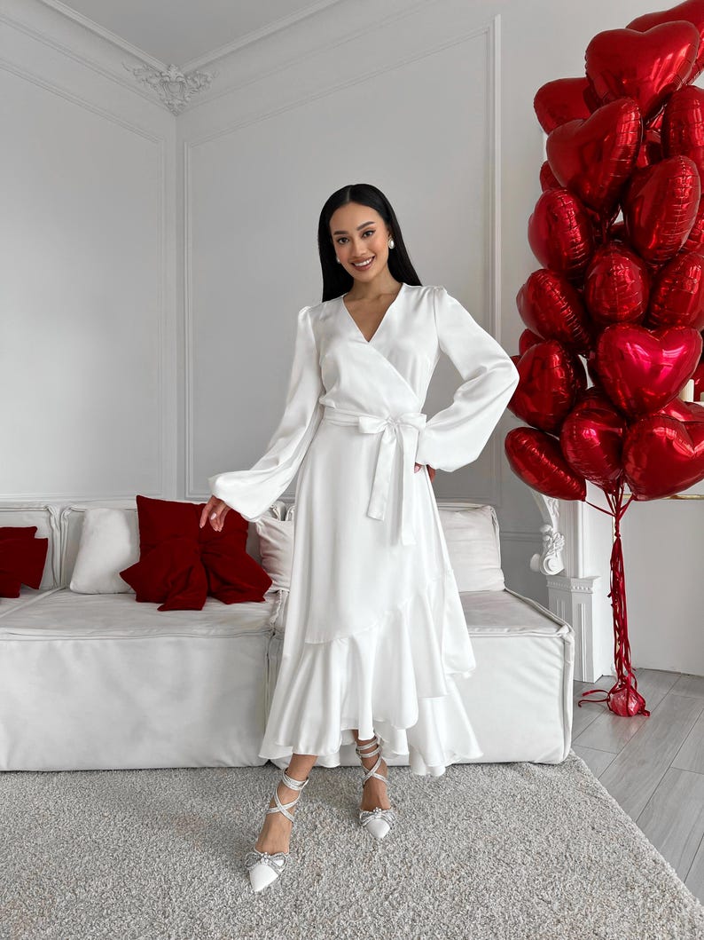 May include: A white wrap dress with long sleeves and a ruffled hem. The dress is tied at the waist with a matching belt. The woman is wearing white heels with bow details. A bouquet of red heart-shaped balloons is in the background.