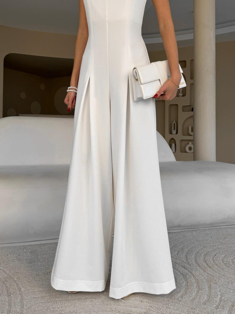 May include: A sleeveless, white jumpsuit with wide legs. The outfit is paired with a small, white clutch. The model is wearing a pearl bracelet. The jumpsuit has a tailored fit and a minimalist design, suitable for formal occasions.