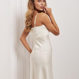 May include: Cream-coloured slip dress with thin straps. The dress features a fitted bodice and a flowing skirt. The model is wearing the dress and has long, wavy blonde hair. The background is a plain white wall.