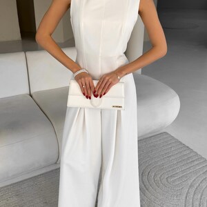 May include: A sleeveless, white jumpsuit with wide legs. The jumpsuit features a high neckline and a fitted bodice. A white clutch with the gold text "JACQUEMUS" is held in front. The outfit is accessorized with a bracelet and rings.