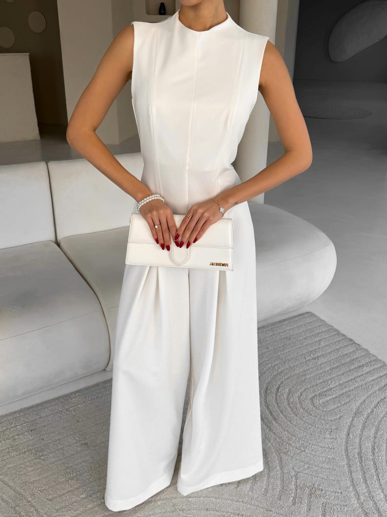May include: A sleeveless, white jumpsuit with wide legs. The jumpsuit features a high neckline and a fitted bodice. A white clutch with the gold text "JACQUEMUS" is held in front. The outfit is accessorized with a bracelet and rings.