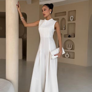 May include: A sleeveless, white jumpsuit with wide legs and a high neckline. The outfit includes a matching white clutch. The model is wearing pearl earrings and a bracelet. The background is a neutral color.
