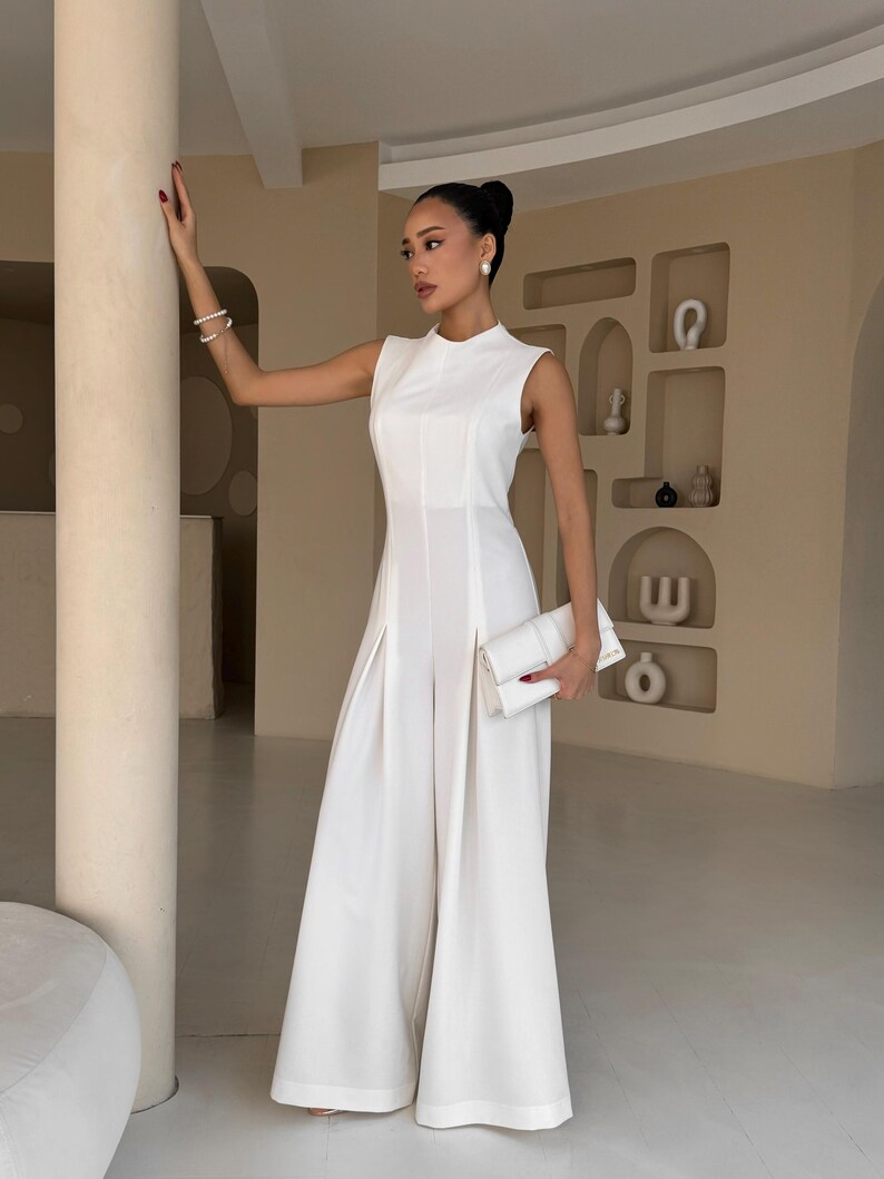 May include: A sleeveless, white jumpsuit with wide legs and a high neckline. The outfit includes a matching white clutch. The model is wearing pearl earrings and a bracelet. The background is a neutral color.