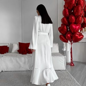 May include: A white maxi dress with a belted waist and a flared hemline. The dress features long sleeves and is worn with white heels. A cluster of red heart-shaped balloons is in the background, alongside a white sofa and red cushions.