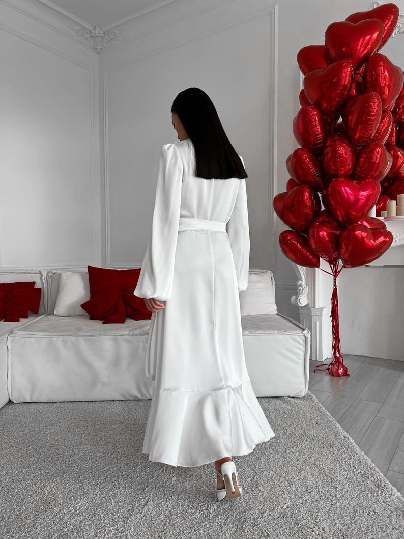 May include: A white maxi dress with a belted waist and a flared hemline. The dress features long sleeves and is worn with white heels. A cluster of red heart-shaped balloons is in the background, alongside a white sofa and red cushions.