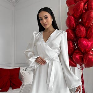 May include: A white satin wrap dress with long sleeves and a tie waist. The dress features a V-neck and a ruffled hem. A bunch of red heart-shaped balloons is in the background.