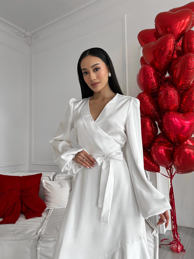 May include: A white satin wrap dress with long sleeves and a tie waist. The dress features a V-neck and a ruffled hem. A bunch of red heart-shaped balloons is in the background.